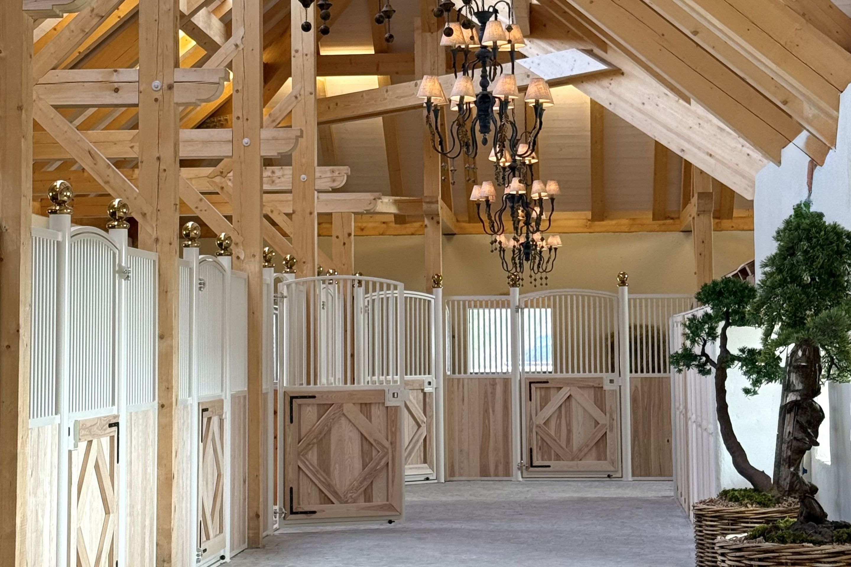 Innenansicht des Hofstalls Schloss Amerang in Bayern mit hellem Holz-Dachstuhl, weißen Pferdeboxen und eleganten Kronleuchtern.